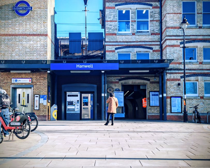 A woman standing outside the entrance of Hanwell station, part of the Elizabeth Line, with a brick building facade featuring multiple windows in the background. The station signage is prominently disp